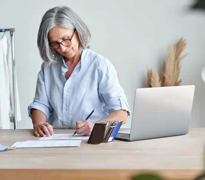 Mature female business owner with gray hair sketching designs at a desk beside a laptop.
